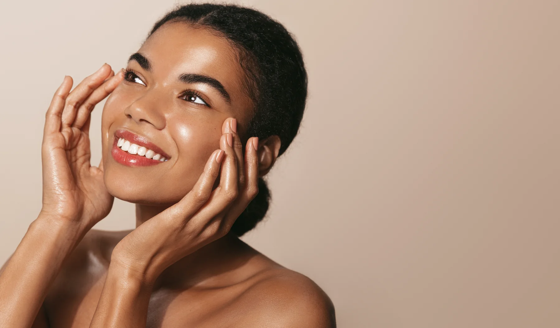 Young african american girl with clear, glowing skin, smiling and looking up as she touches her smooth skin after a facial.